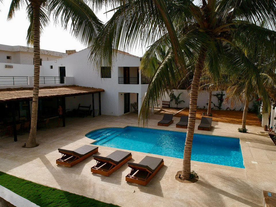 Piscine rectangulaire bordée de transats, devant une villa à étage et des palmiers